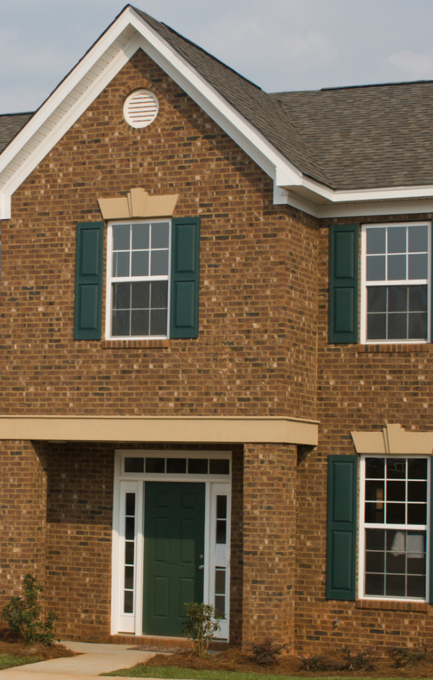 Two-story brick house with green shutters and a green front door, small bushes in front.